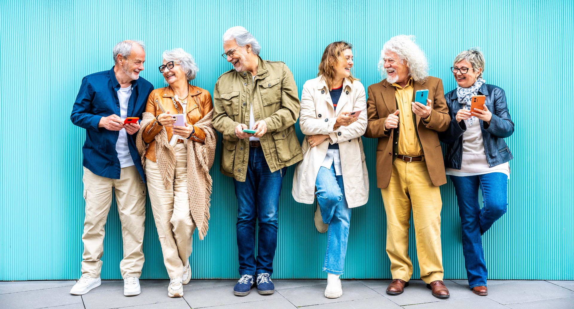Gruppo di persone anziane che utilizzano dispositivi tecnologici insieme in piedi su una parete blu - Amici anziani felici che si divertono a guardare video divertenti su smartphone - Concetto di anziani tecnologici e moderni